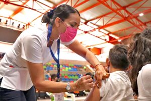 Enfermera del sector de salud aplicando vacuna contra covid-19 a menor de edad en escuela primaria.
